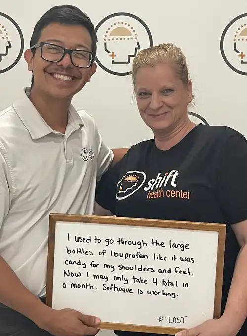 Two people stand side by side in front of a Shift Health Center backdrop, holding a whiteboard testimonial. The whiteboard shares a patient story about significantly reducing ibuprofen use for shoulder and foot discomfort after treatment.